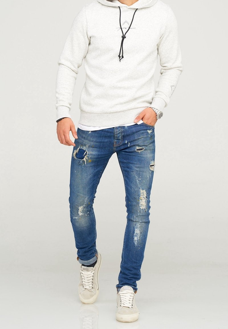 Hellgrau Hoodie mit schwarzen Kordeln, kombiniert mit durchgewetzten blauen Jeans mit Rissen und Farbspritzern, getragen mit beigen Turnschuhen.