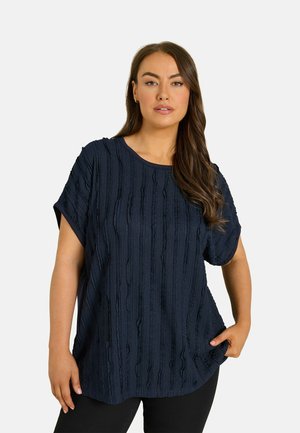 Vrouw met lang bruin haar, gekleed in een gestructureerde donkerblauwe top met korte mouwen en zwarte broek, staand met één hand in haar zak, neutrale achtergrond.