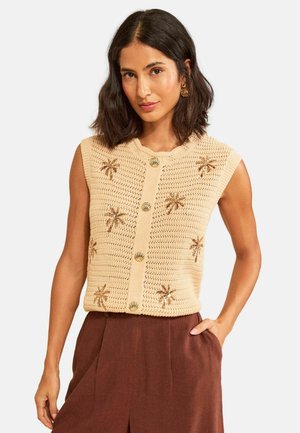 Frau trägt eine beige ärmellose Strickweste mit Palmenmustern und Muschelknoöpfen, kombiniert mit hoch taillierten braunen Hosen.