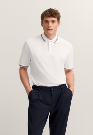 White polo shirt with a black-striped collar, featuring a button placket and short sleeves, worn with navy trousers. Smooth texture.