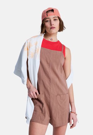 Brązowe lniane ogrodniczki z szortami, z dwoma przednimi kieszeniami, nałożone na czerwony top. Biała kurtka z delikatnym wzorem spoczywa na ramieniu. Różowa czapka.