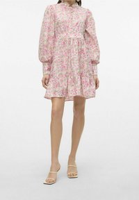 Kvinna iklädd en rosa och vit blommönstrad mini-klänning med puffade långärmar och beige öppetåad fyrkantiga klackskor, stående mot en vit bakgrund.