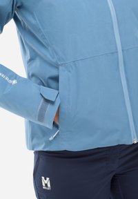 Veste imperméable bleu clair avec fermeture éclair à l'avant, poignets ajustables avec Velcro et design de poche dissimulée. Logo affiché sur le côté.