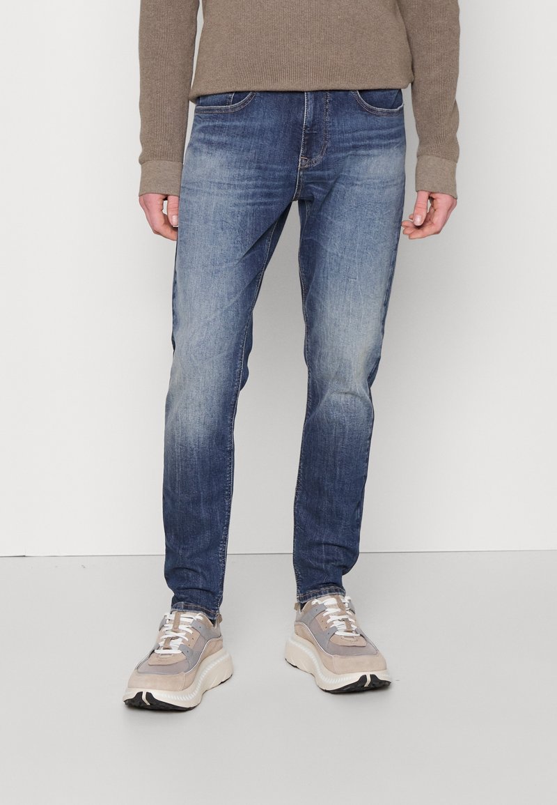 Person, der verblasste blaue Jeans, einen beigefarbenen Pullover und beigefarbene und graue klobige Sneakers trägt, steht vor einem schlichten weißen Hintergrund.