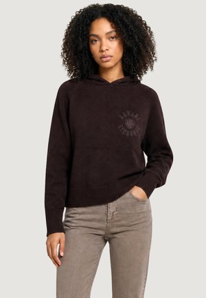 Kobieta z kręconymi włosami nosi ciemnobrązową bluzę z kapturem z logo Armani Exchange oraz jasnobrązowe jeansy, jedna ręka w kieszeni, neutralne tło.