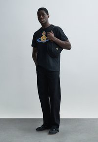 Μαύρο μπλουζάκι με γραφικό σχέδιο, μαύρο παντελόνι και μαύρα σανδάλια. Μια καμπυλωτή μαύρη τσάντα με φερμουάρ φοριέται στον ώμο.