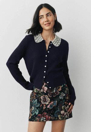 Marineblaue gerippte Strickjacke mit einem Perlenkragen, ausgestattet mit Perlenknöpfen an der Vorderseite. Kombiniert mit einem blumengemusterten Rock in lebhaften Farben.