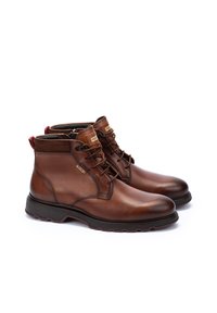 Botas de tobillo de cuero marrón con un acabado suave, que cuentan con un diseño de cordones, una lengüeta de agarre roja en contraste y una suela de goma para mayor tracción y durabilidad.