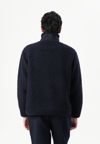Geacă din fleece bleumarin, cu un exterior texturat și pufos, guler înalt și croială lejeră, concepută pentru căldură și confort.