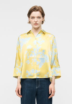 Lühikeste juustega naine, kes kannab lahtist kollast nööpidega särki helesiniste lillemustritega ja tumesiniseid teksaseid, seistes ühevärvilise tausta ees.