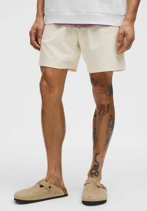 Mand iført cremefarvede shorts og beige slip-on sko, der viser flere tatoveringer på højre ben fra lår til ankel.