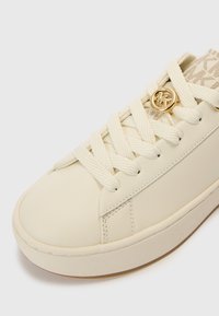 Kremaste tenisice s glatkim sintetičkim gornjim dijelom, okruglim vrhom, debelim pertlama i zlatnim detaljem s logotipom. Teksturirani tkaninski naglasci na jezičku.