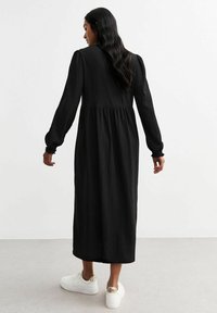Vestido negro de manga larga con tejido texturizado, cintura fruncida y falda fluida. Llevado con zapatillas blancas con acentos dorados.