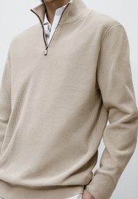 Beiger, gerippter Strickpullover mit einem Viertelreißverschluss-Kragen, der über einem weißen Hemd getragen wird.