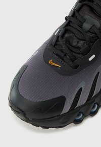 Sivi atletski tenisice s teksturiranim gornjim dijelom, crnim detaljima, mrežastim elementima i zlatnim Nike logom. Imaju okrugli vrh i vidljivo zračno jastučić.