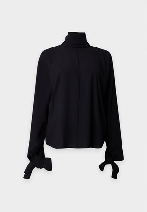 Kvinde med langt sort hår iført en sort rullekravetrøje, blå skinny jeans og sorte spidse flats, der står foran en ensfarvet baggrund.