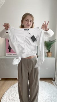 Balta ilga rankovė marškiniai su etikete, pasižymintys laisvu siluetu ir lygia tekstūra. Modelis laiko juos prieš neutralų foną.