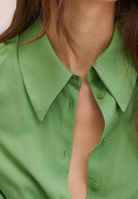 Camicia verde con colletto grande, texture liscia e dettagli di cucitura sottili. Presenta due bottoni vicino allo scollo.