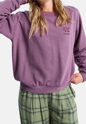 Donna che indossa una felpa viola Roxy e pantaloni verdi a quadri, posando con una mano dietro la testa e l'altra in tasca.