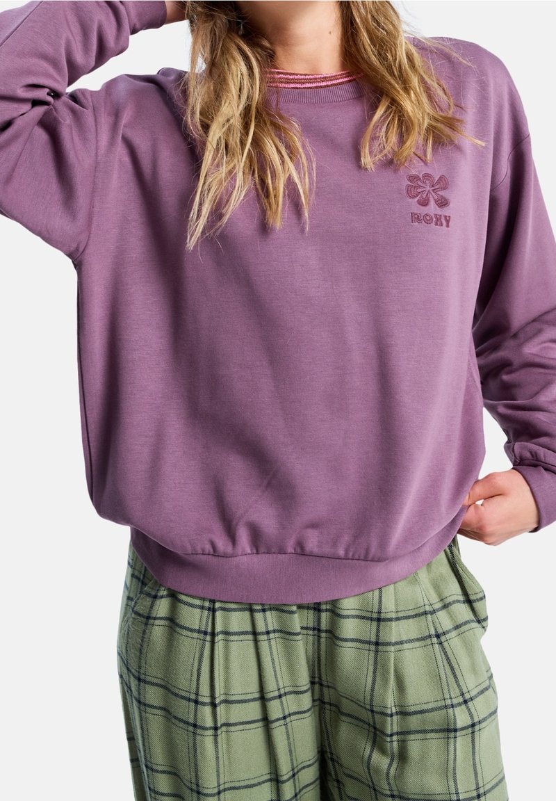 Femme portant un sweat-shirt violet Roxy et un pantalon vert à carreaux, posant avec une main derrière la tête et l'autre dans la poche.