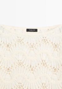 Witte kanten stof met bloem- en geometrische haakpatronen en een zwart label met de tekst "Massimo Dutti Made in China" bij de halslijn.