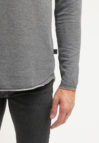 Maglietta a maniche lunghe grigio scuro con finitura testurizzata, caratterizzata da orli grezzi e un'etichetta sottile di marca sul lato. Indossata con jeans scuri.