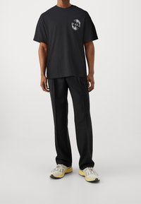 Svart bomullströja med grafisk design på vänster bröst, kombinerad med svarta byxor och flerfärgad sportskor.