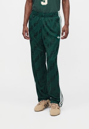 Mand iført mørkegrønne Adidas træningsbukser med hvide striber ned langs siderne og beige sneakers, stående foran en ensfarvet baggrund.