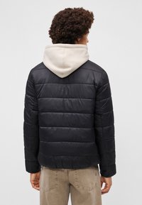 Melnā pūkainā jaka ar horizontālu quilted dizainu, zem tās ir bežs kapuci, izgatavota no gludas, ūdensizturīgas materiāla.