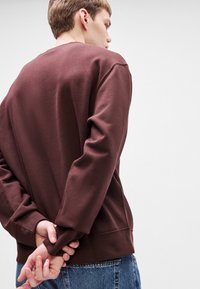 Καφετί φούτερ με μακριά μανίκια, μαλακή υφή, σχεδίαση με στρογγυλή λαιμόκοψη και ραφές, συνδυασμένο με μπλε τζιν παντελόνι.