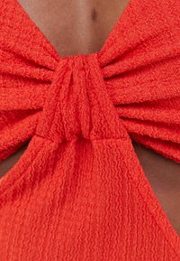 Červená texturovaná tkanina se vrstveným designem mašle uprostřed, obsahující sklady, které vytvářejí vlnitý vzhled, a hladkou podšívku.