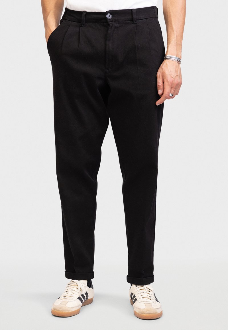 Black tapered trousers made from smooth material, featuring front pleats, belt loops, and side pockets. Paired with light sneakers.