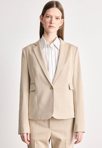 Bežový blazer s textúrovanou tkaninou, zapínanie na jeden gombík a dve predné vrecká. V kombinácii s bielou košeľou s golierom a prúžkami pod ním.