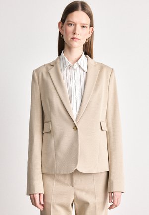 Blazer beige con tejido texturizado, cierre de un botón y dos bolsillos frontales. Combinado con una camisa blanca de rayas con cuello debajo.
