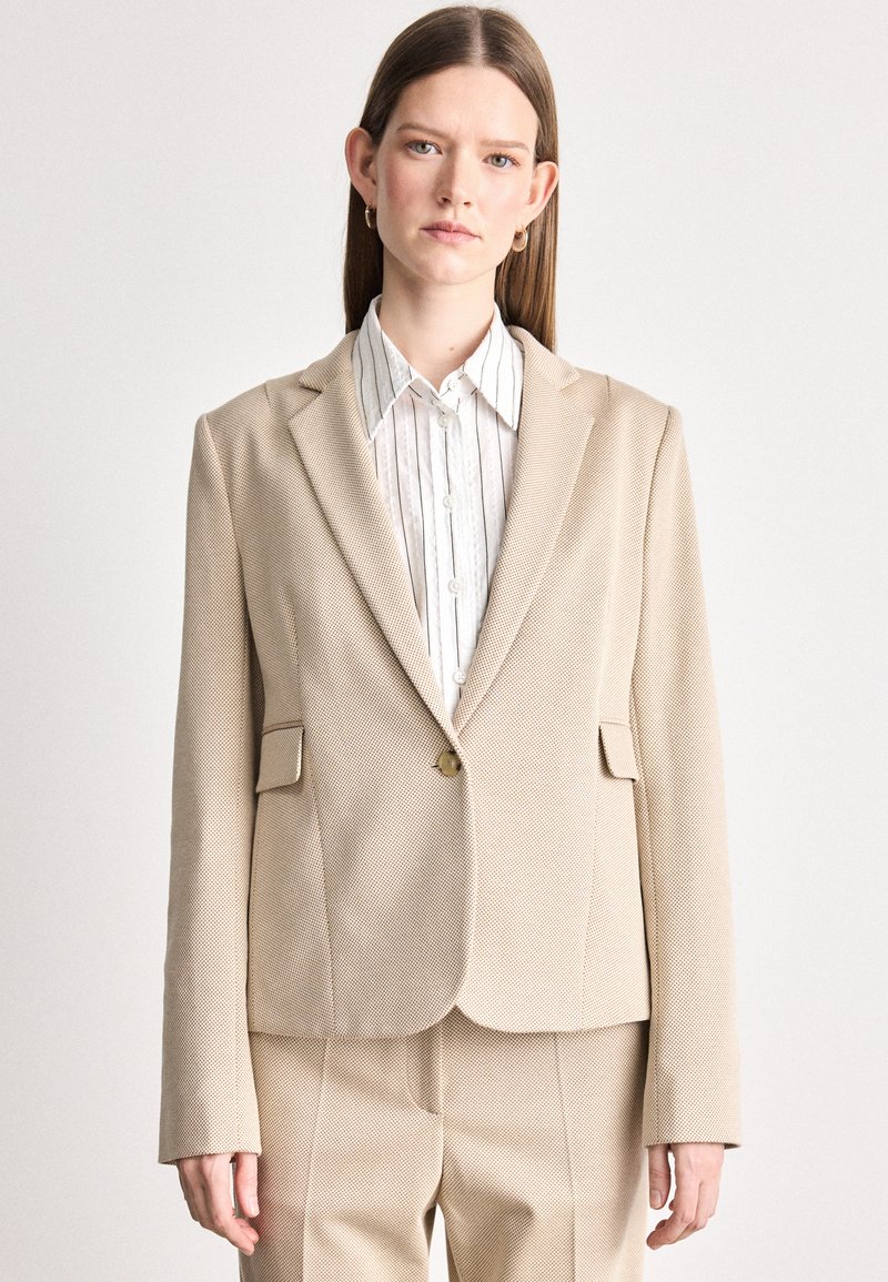 Bežový blazer s textúrovanou tkaninou, zapínanie na jeden gombík a dve predné vrecká. V kombinácii s bielou košeľou s golierom a prúžkami pod ním.