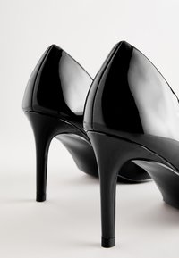 Zapatos de tacón alto de charol negro con puntera afilada, que presentan un diseño elegante y un acabado brillante. Los tacones de aguja tienen aproximadamente 10 centímetros de altura.