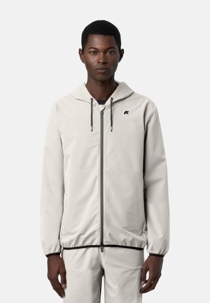 Uomo che indossa una giacca con cappuccio beige chiaro con zip e pantaloni coordinati, in piedi contro uno sfondo bianco uniforme.