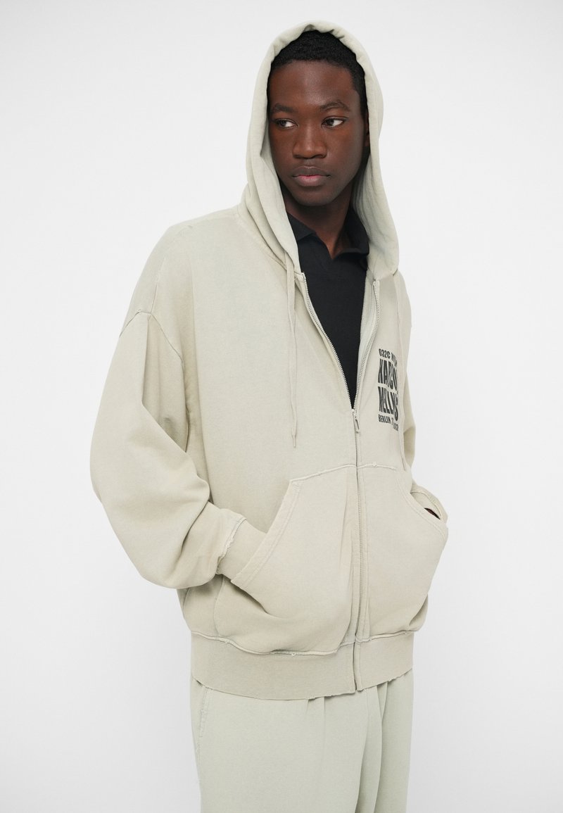 Hoodie zippé beige clair en tissu doux avec une poche avant, coupe oversize et capuche, orné de graphiques minimalistes.