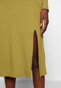 Olivengrünes, geripptes Kleid mit Seitenschlitz, langen Ärmeln und einer glatten, weichen Textur. Ausschnitt und Saum sind sauber verarbeitet.