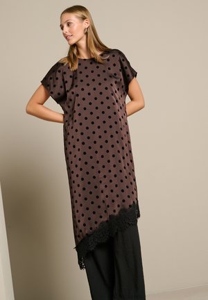 Femme portant une robe marron ample avec des pois noirs et une bordure en dentelle, debout les mains dans le dos sur un fond uni.