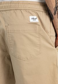 Pantalones cortos beige de mezcla de algodón, con una cintura elástica, bolsillo trasero y una etiqueta cosida con el nombre de la marca en la parte trasera.