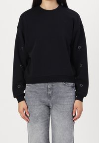 Femme portant un sweat-shirt noir avec de petits cœurs blancs en contour sur les manches et un jean gris clair délavé, sur un fond uni.