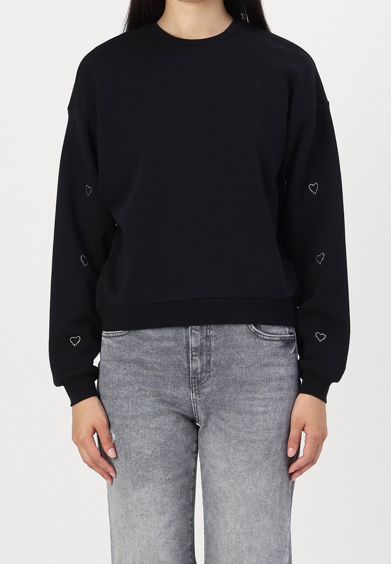Femme portant un sweat-shirt noir avec de petits cœurs blancs en contour sur les manches et un jean gris clair délavé, sur un fond uni.
