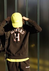 Felpa nera con testo "HARVARD" in bianco e rosso; cappellino giallo; zaino nero; maglietta gialla; pantaloni della tuta con dettagli del logo.