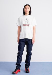 Witte katoenen T-shirt met een grafisch ontwerp featuring figuren en de tekst "FAIRPLAY", gecombineerd met donkere denim jeans en zwart/rode sneakers.