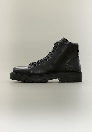 Bottine noire en cuir à lacets, dotée d’une épaisse semelle plateforme, présentée de profil sur un fond uni.