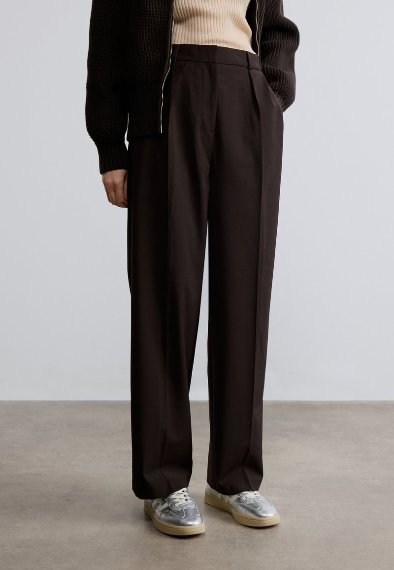 Pantalon large marron foncé en tissu léger, présentant des plis et une texture lisse ; assorti de baskets grises et d'un pull en maille noir.