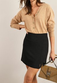 Vrouw in een beige geknoopte trui en zwarte rok met zijsplit, die een gestructureerde beige handtas met metalen schouderband vasthoudt.