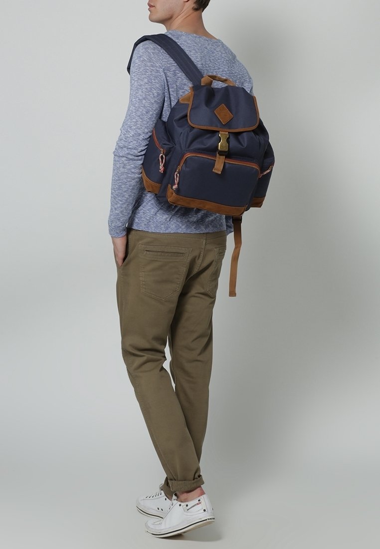 Homme portant une chemise à manches longues bleue, un pantalon beige, des baskets blanches, portant un sac à dos bleu marine et marron, se tenant devant un fond gris uni.