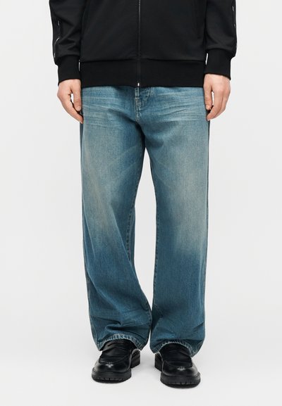 Άνδρας που φοράει ξεθωριασμένο, άνετο μπλε τζιν παντελόνι, μαύρα παπούτσια και μαύρο μπουφάν με φερμουάρ, στέκεται μπροστά σε απλό φόντο.